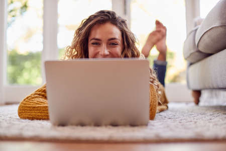 Woman In Warm Jumper Lying On Floor At Home Using Laptop