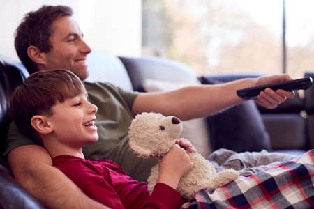 Father And Son With Toy Dog Sitting On Sofa In Pyjamas Together Watching TV