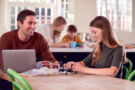 Father Helps Teenage Daughter With Electronics Project Sitting At Kitchen Table At Home With Laptop