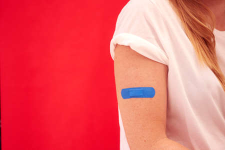 Woman With Blue Sticking Plaster On Arm After Injection Or Vaccination Against Red Backgroundの写真素材