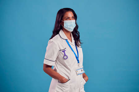 Portrait Of Female Mature Nurse In Uniform Wearing Face Mask In Front Of Blue Studio Backgroundの写真素材