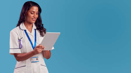 Smiling Female Mature Nurse Wearing Uniform With Digital Tablet In Front Of Blue Studio Background