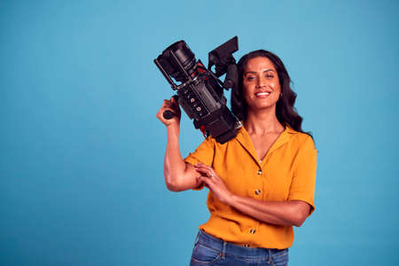 Portrait Of Mature Female Videographer With Camera Filming Video Against Blue Background In Studioの写真素材