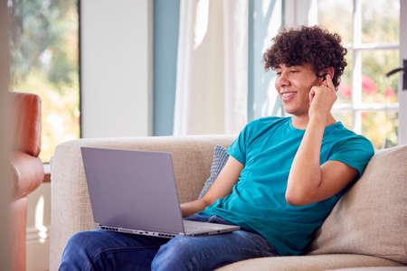 Teenage Boy Sitting On Sofa At Home Using Laptop Computer And Talking On Mobile Phone