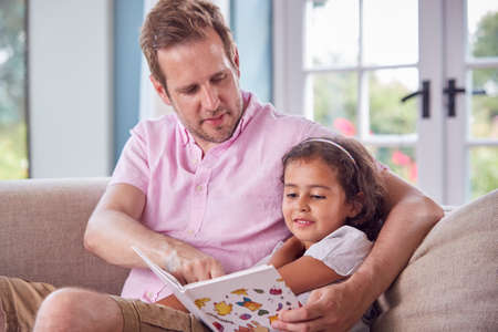 Father And Daughter Sitting On Sofa At Home Reading Book Together
