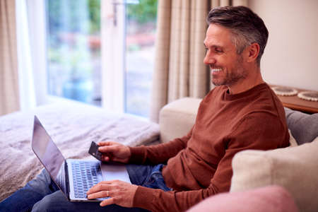 Mature Man At Home Sitting On Sofa With Laptop Paying For Online Purchase With Credit Card