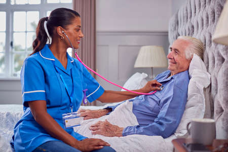 Female Care Worker In Uniform Listening To Chest Of Senior Man At Home In Bed With Stethoscopeの写真素材