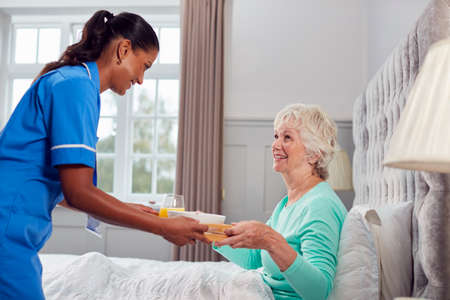 Female Care Worker In Uniform Bringing Senior Woman At Home Breakfast In Bed On Trayの写真素材