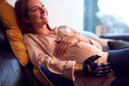 Close Up Of Pregnant Woman With Prosthetic Arm Relaxing Lying On Sofa At Home Holding Bumpの写真素材
