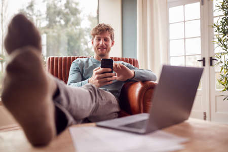 Man Relaxing In Armchair With Feet Up At Home Using Laptop And Mobile Phoneの写真素材