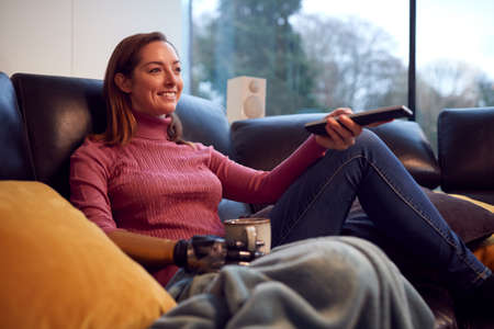 Woman With Prosthetic Arm And Hand At Home Watching TV And Drinking Cup Of Coffee