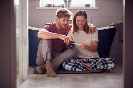 Excited Couple With Woman With Prosthetic Arm Sitting On Bathroom Floor With Positive Pregnancy Test