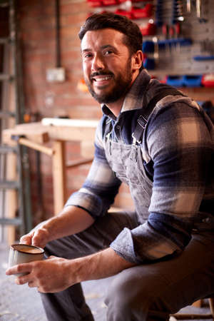 Portrait Of Smiling Man Wearing Overalls In Garage Workshop With Hot Drinkの写真素材