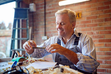 Mature Male Carpenter In Garage Workshop Taking Photo Of Design On Mobile Phoneの写真素材