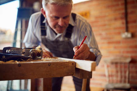 Mature Male Carpenter In Garage Workshop Marking Wood With Pencil