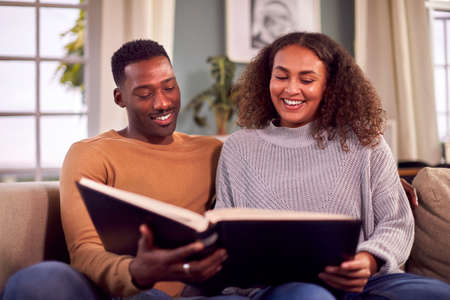 Smiling Couple Sitting On Sofa At Home Looking Through Photo Albumの写真素材