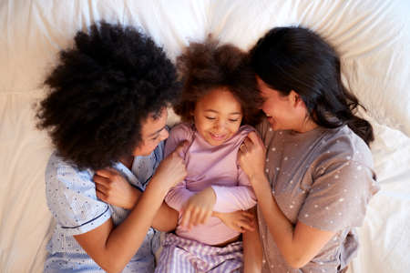 Overhead Shot Of Family With Two Mums Wearing Pyjamas Playing On Bed At Home With Daughterの写真素材