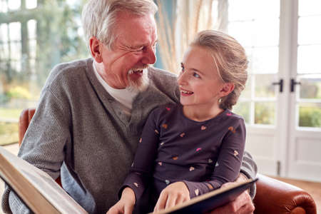 Grandfather And Granddaughter Looking Through Photo Album In Lounge At Home Together