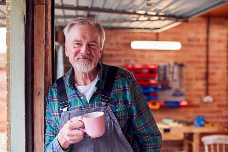 Portrait Of Senior Male Wearing Overalls In Garage Workshop With Hot Drink