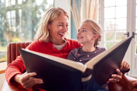 Grandmother And Granddaughter Looking Through Photo Album In Lounge At Home Together