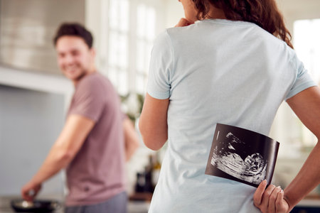 Pregnant Transgender Couple At Home In Kitchen With Woman Surprising Man With Ultrasound Scan Of Babyの写真素材