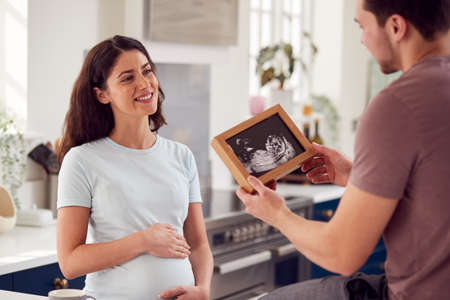 Excited Pregnant Transgender Couple At Home In Kitchen Looking At Ultrasound Scan Of Babyの写真素材