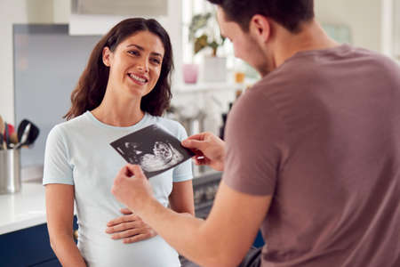 Excited Pregnant Transgender Couple At Home In Kitchen Looking At Ultrasound Scan Of Babyの写真素材