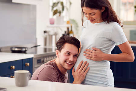 Excited Pregnant Transgender Couple At Home In Kitchen With Man Listening To Baby's Heartbeatの写真素材
