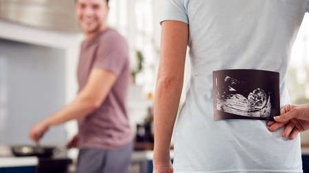 Pregnant Transgender Couple At Home In Kitchen With Woman Surprising Man With Ultrasound Scan Of Babyの写真素材