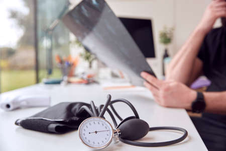 Close Up Of Male Doctor Sitting At Desk With CT Or MRI Scan And Blood Pressure Sphygmomanometer