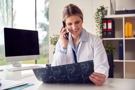 Female Doctor Or GP Wearing White Coat On Phone Sitting At Desk In Office Looking At CT Or MRI Scan