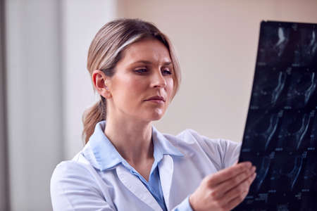 Female Doctor Wearing White Coat Standing In Hospital Corridor Looking At CT Or MRI Scanの写真素材