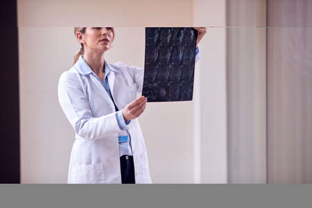 Female Doctor Wearing White Coat Standing In Hospital Corridor Looking At CT Or MRI Scanの写真素材