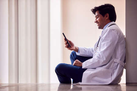 Male Doctor Wearing White Coat Sitting On Floor In Hospital Corridor Text Messaging On Mobile Phoneの写真素材