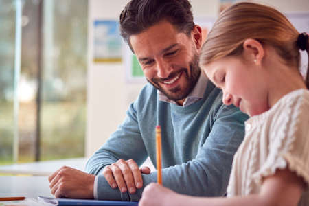 Male Teacher With Female Student In School Classroom Sitting At Desk Writing In Book Togetherの写真素材