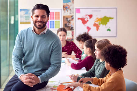 Portrait Of Smiling Male Elementary School Teacher Working At Desk In Classroom With Studentsの写真素材