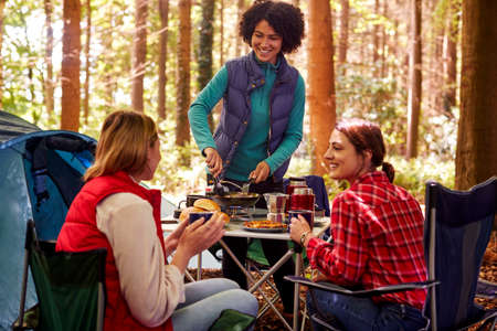 Group Of Female Friends On Camping Holiday In Forest Eating Meal Sitting By Tent Togetherの写真素材