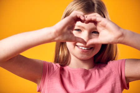 Studio Portrait Of Smiling Girl Making Heart Shape With Hands Against Yellow Backgroundの写真素材