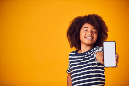 Studio Portrait Of Boy Showing Mobile Phone With Social Media Screen Against Yellow Backgroundの写真素材