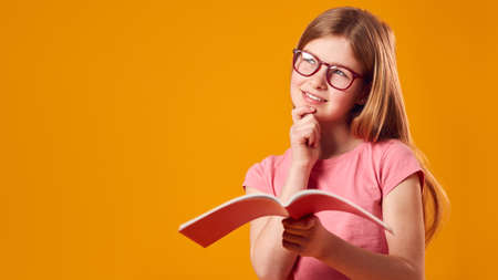 Studio Shot Of Young Girl Studying School Exercise Book Wearing Glasses Against Yellow Backgroundの写真素材