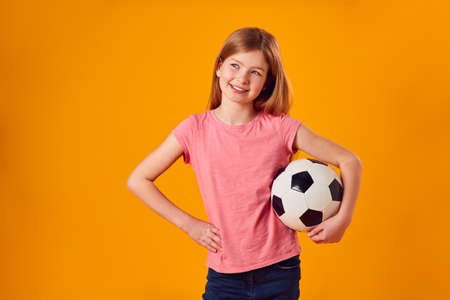 Studio Shot Of Young Girl Holding Soccer Ball Under Arm Against Yellow Backgroundの写真素材