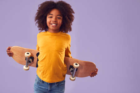 Studio Portrait Of Young Boy Holding Skateboard Behind Back Against Purple Background