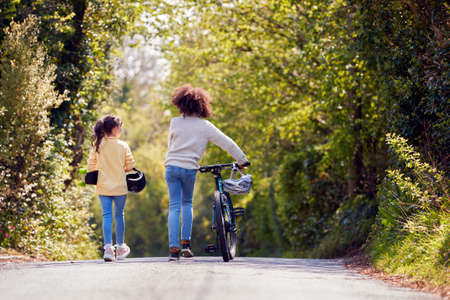 Rear View Of Boy With Bike And Girl With Skateboard Walking Along Country Road Together