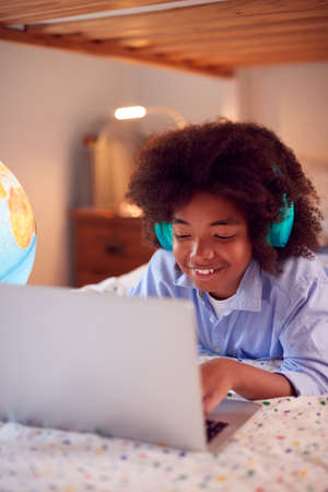Boy In Bedroom At Night Lying On Bed Using With Laptop With Illuminated Globe