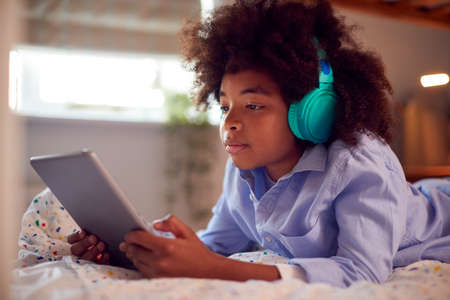 Boy In Bedroom Lying On Bed Wearing Headphones Using Digital Tablet