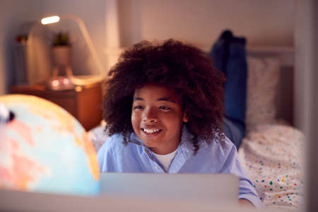 Boy In Bedroom At Night Lying On Bed Using With Laptop With Illuminated Globe