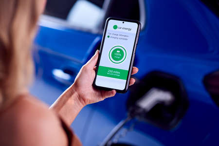 Woman Charging Electric Car With Cable Using App On Phone To Monitor Battery Level