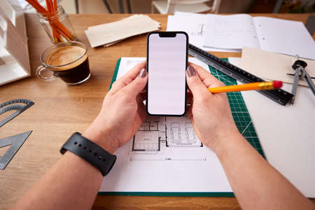 POV Shot Of Female Architect Working In Office On Plans For New Building Using Mobile Phoneの写真素材