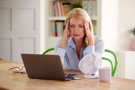 Menopausal Mature Woman Having Hot Flush At Home Cooling Herself With Fan Connected To Laptopの写真素材
