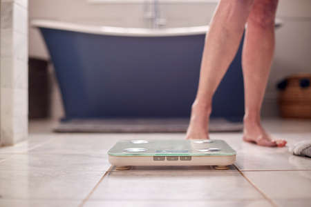 Close Up Of Menopausal Mature Woman Concerned With Weight Gain Standing On Scales In Bathroomの写真素材